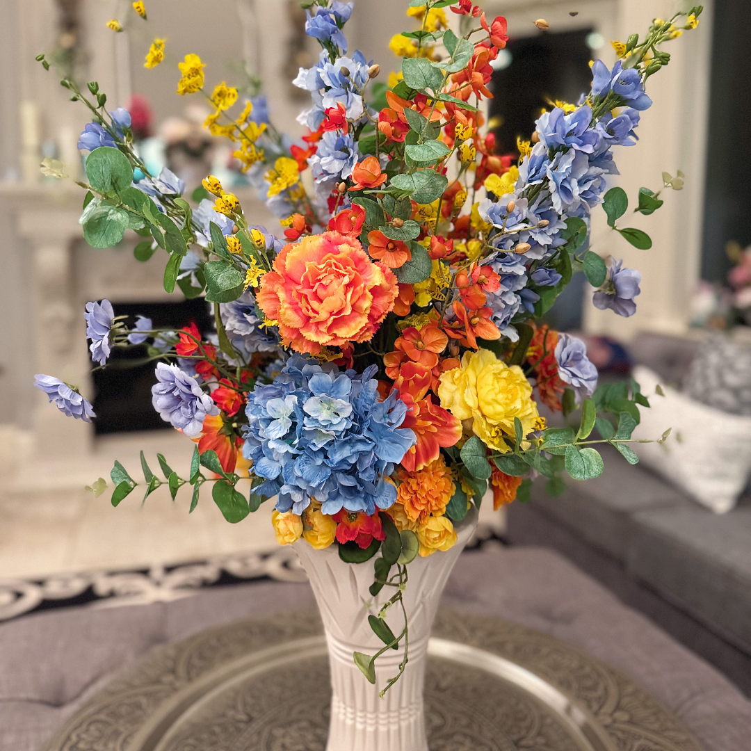 Sunset Meadow Bouquet Vase arrangement featuring soft blue delphiniums, a blue hydrangea, coral and orange carnations, golden yellow blooms, airy yellow spray flowers, and eucalyptus in a white sculpted vase, styled on a coffee table with a softly blurred living room background.