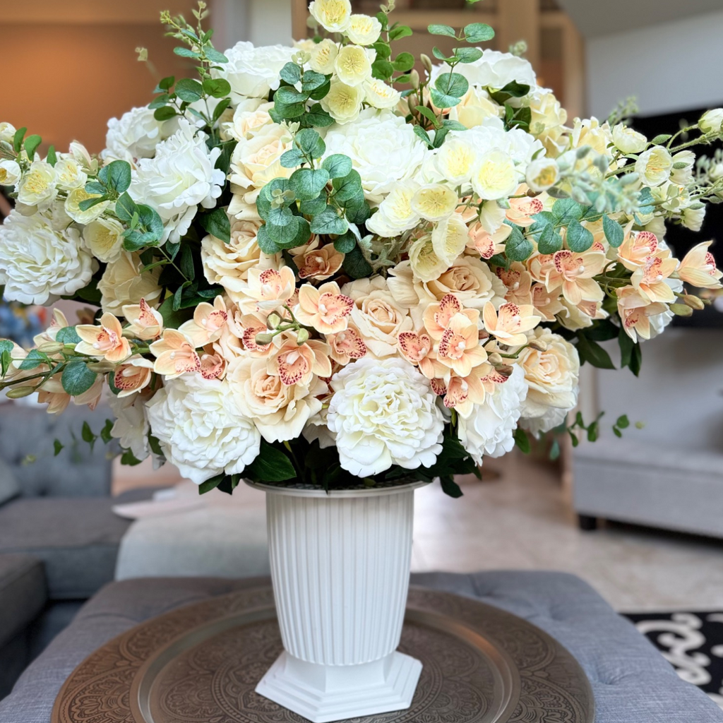 Grand Palais Premium Collection permanent floral arrangement in the Peaches & Cream palette featuring layered ivory peonies, cream roses, peach cymbidium orchids, creamy delphiniums, and eucalyptus in a white Grand Palais Bowl.