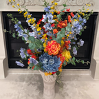 Sunset Meadow Bouquet Vase arrangement featuring soft blue delphiniums, a blue hydrangea, coral and orange carnations, golden yellow blooms, airy yellow spray flowers, and eucalyptus in a white sculpted vase, styled in front of an elegant stone fireplace.