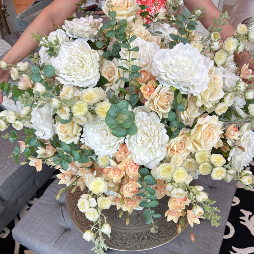 Overhead view of a Grand Palais Premium Collection permanent floral arrangement in the Peaches & Cream palette featuring layered ivory peonies, cream roses, peach cymbidium orchids, creamy delphiniums, and eucalyptus in a white Grand Palais Bowl.