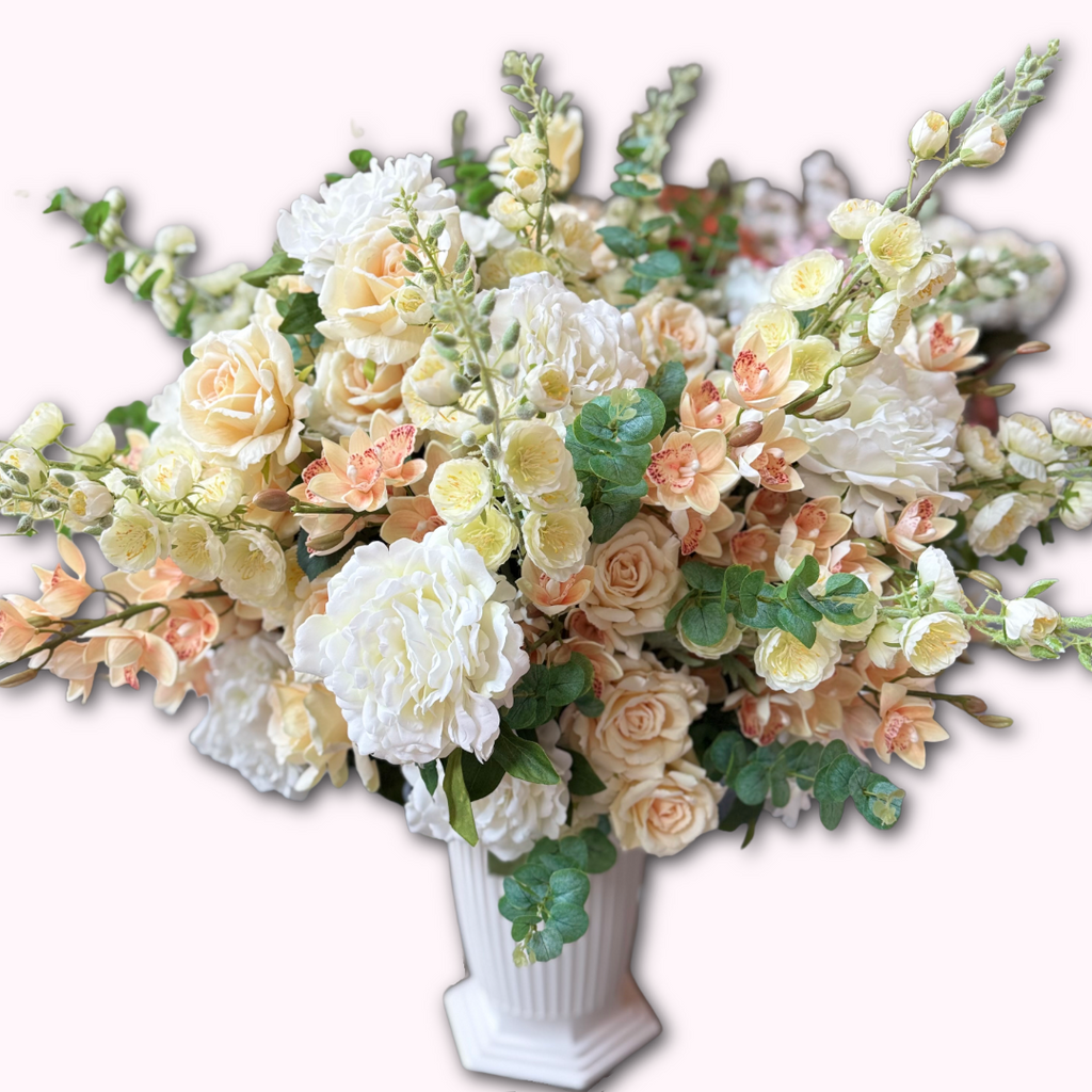 Grand Palais Premium permanent floral arrangement in the Peaches & Cream palette displayed in a white Grand Palais Bowl, featuring lifelike ivory peonies, cream roses, peach cymbidium orchids, creamy delphiniums, and eucalyptus.