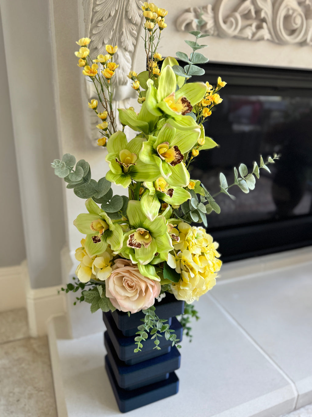 Floral arrangement with green orchids, yellow flowers, and pink roses in a dark blue vase on a white surface.