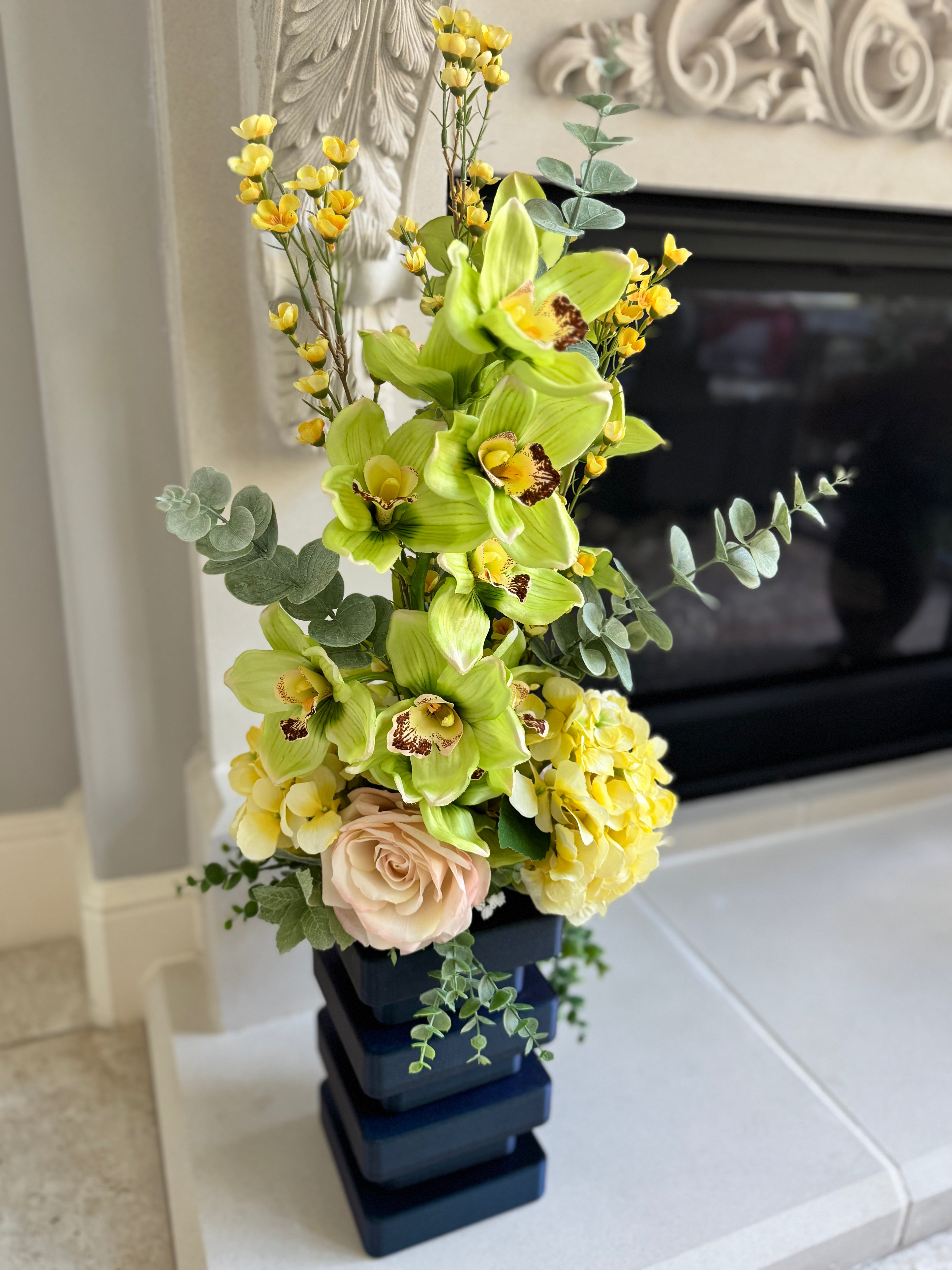Floral arrangement with green orchids, yellow flowers, and pink roses in a dark blue vase on a white surface.