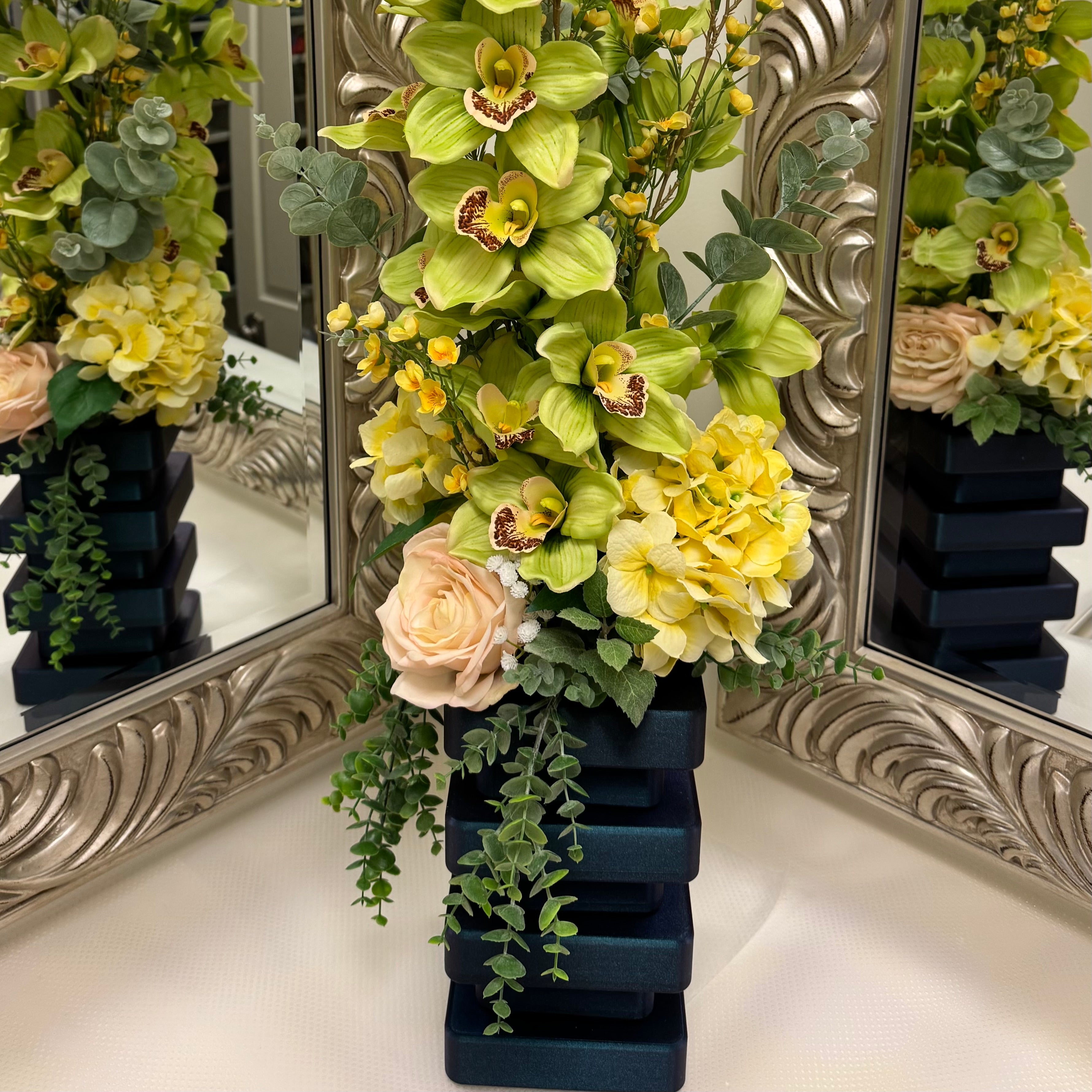 Floral arrangement with green orchids and yellow flowers in a dark blue vase, reflected in ornate mirrors.