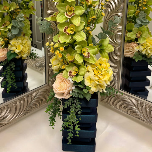 Floral arrangement with green orchids and yellow flowers in a dark blue vase, reflected in ornate mirrors.