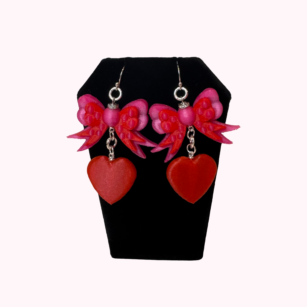 Bow & Heart Dangle Earrings with layered pink bows, red heart charms, and genuine 925 sterling silver hooks displayed on a black jewelry stand.